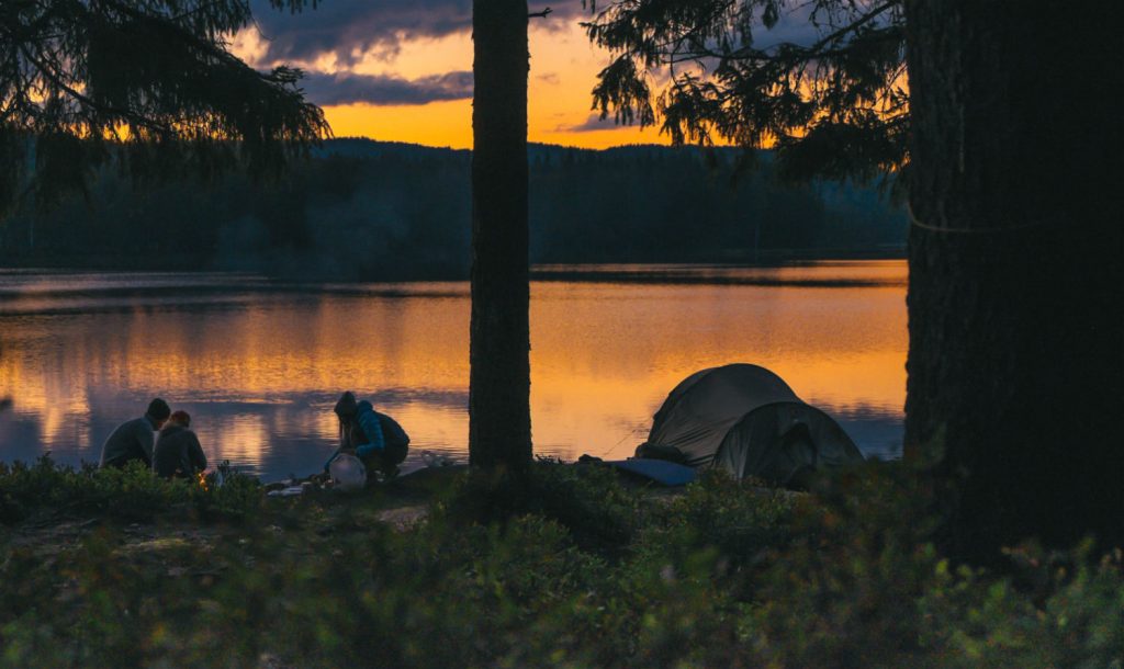 Group of campers making a fire beside their tent along a lake.