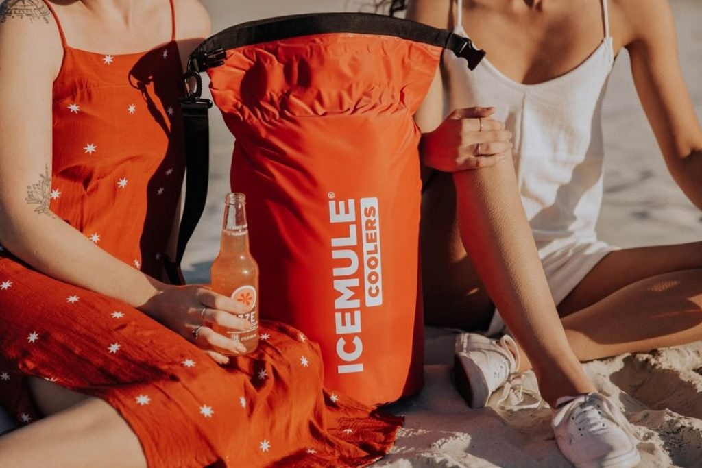 an IceMule cooler in orange in between two women drinking a soda