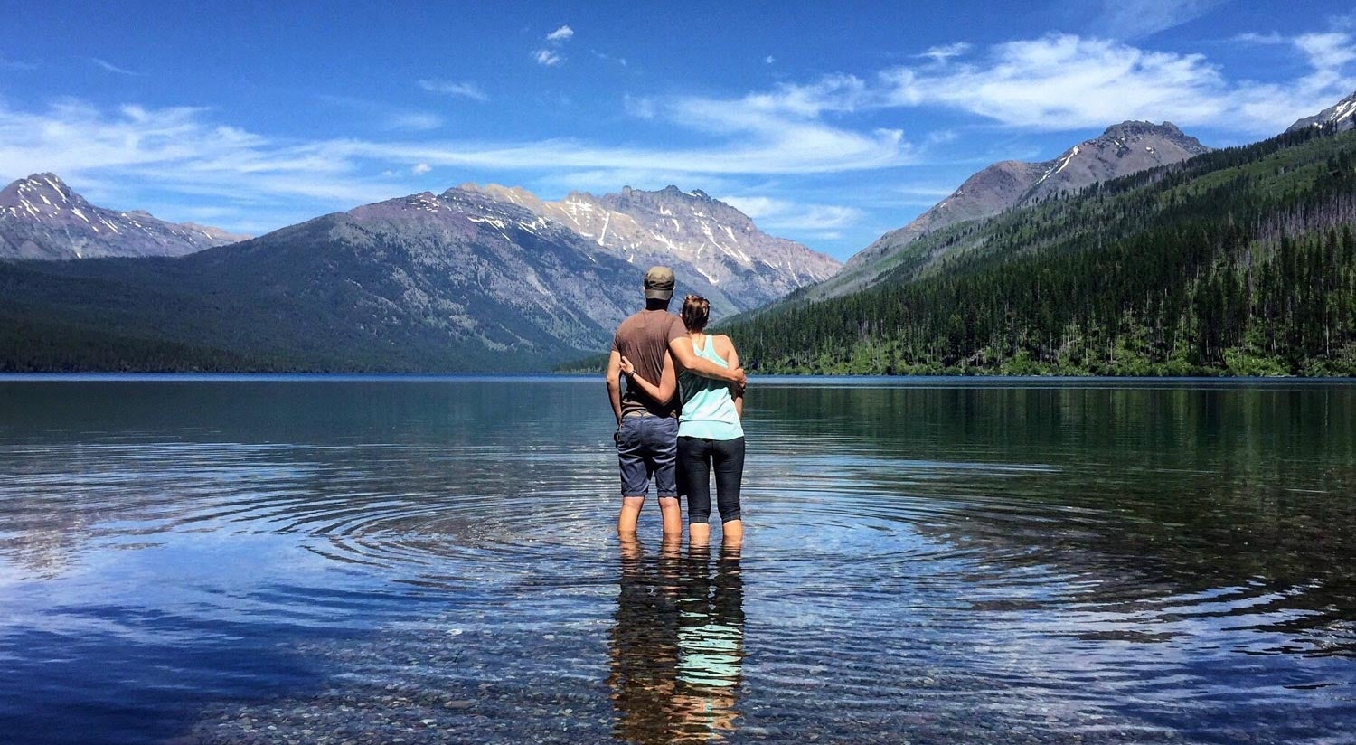 a couple wading in bowman lake embrace and face mountain views as water ripples at their feet