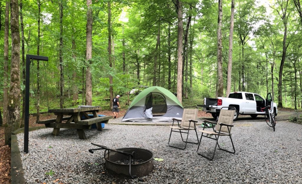 green tent rests in the middle of wooded campsite with picnic table and fire rings
