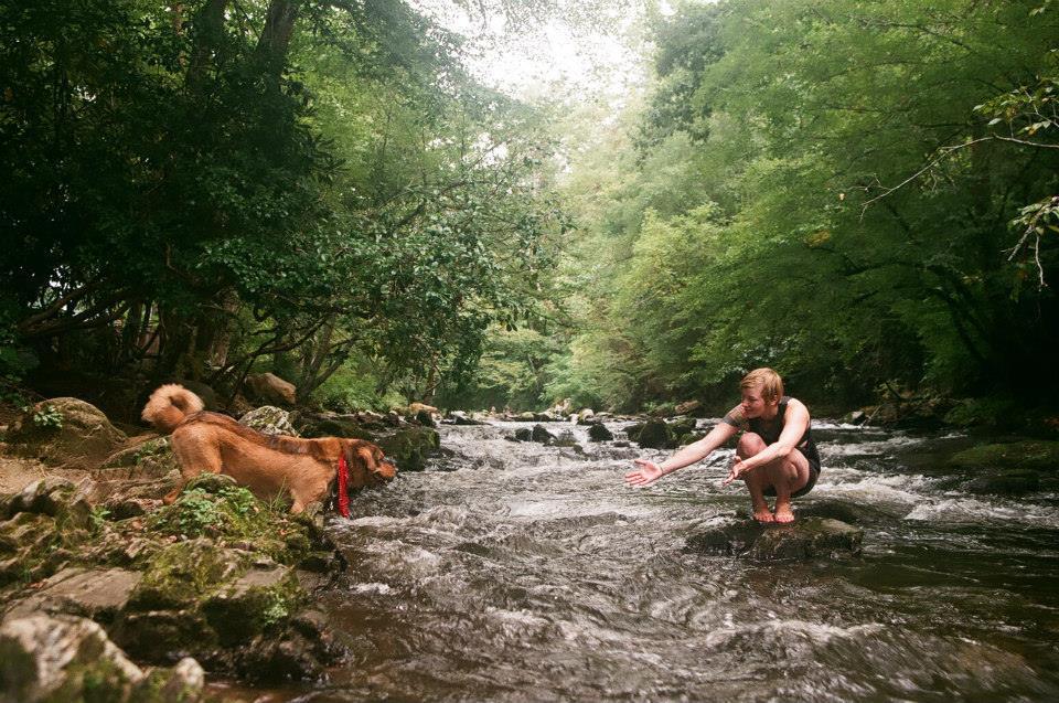woman kneels on a rock in a river as she holds her arms out for a running dog