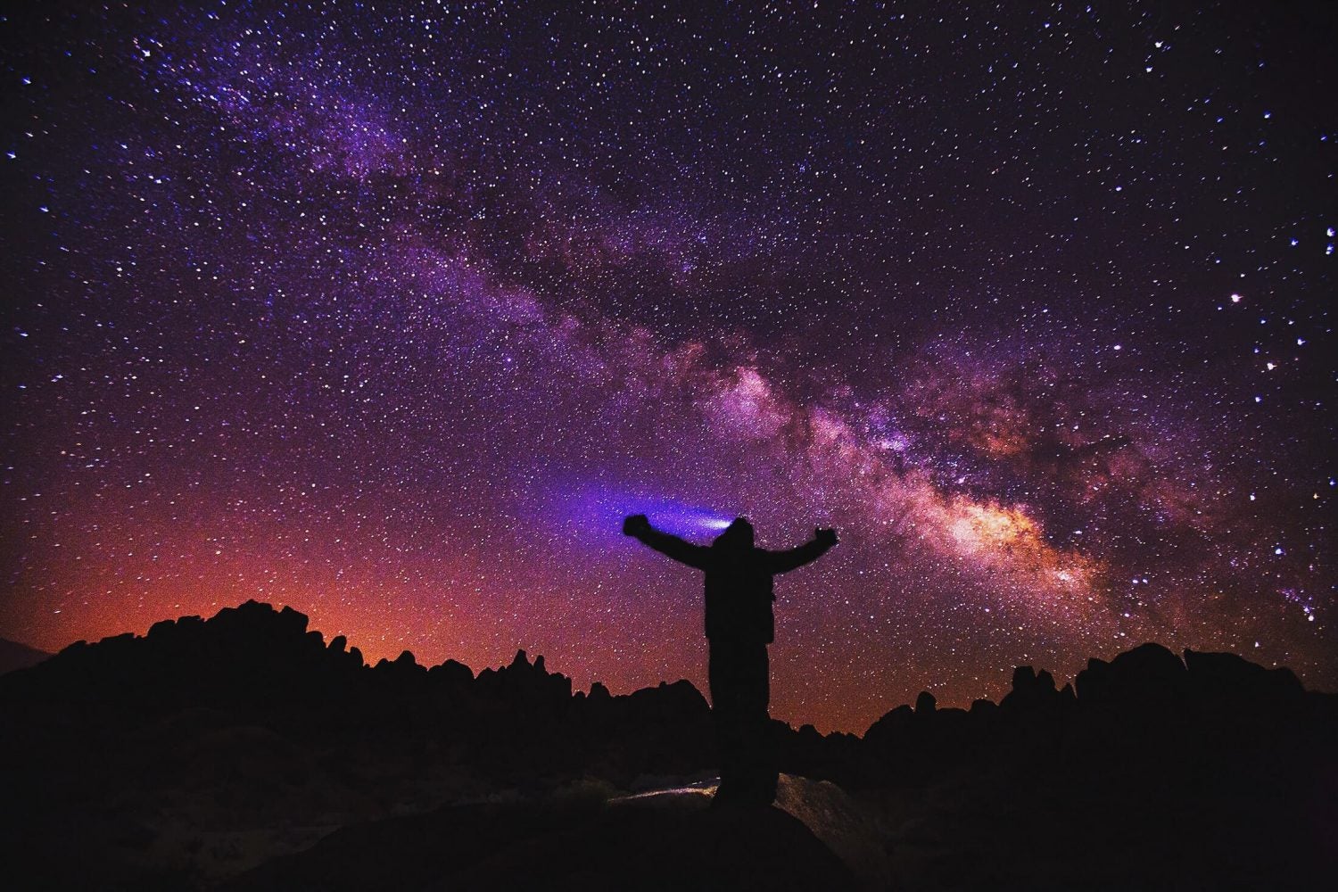 silhouette of a hiker wearing a headlamp as milky way is seen above in the purple and pink night sky