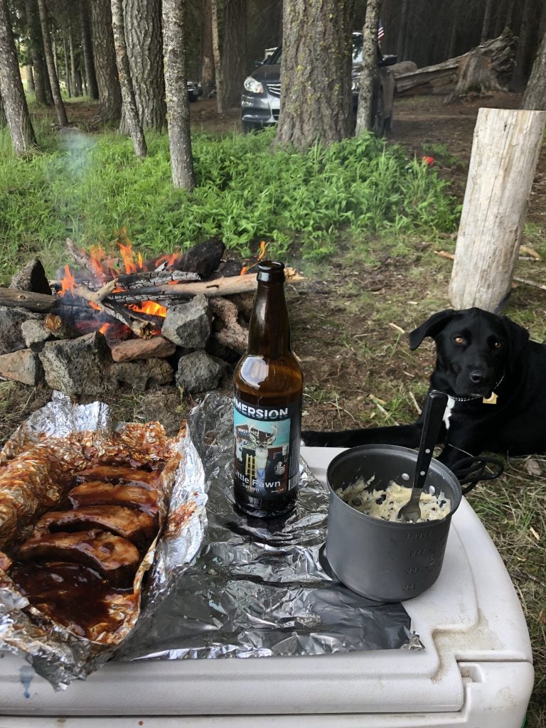 Dog sits among cooked food and a beer around a campfire. 