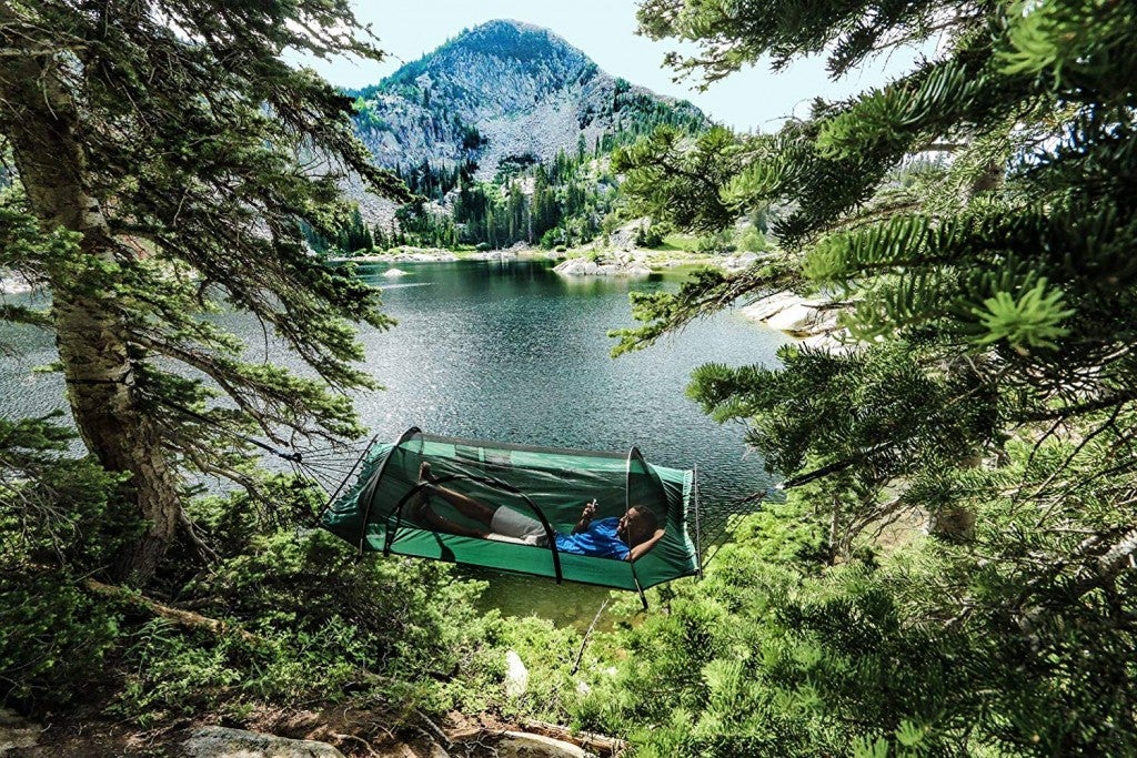 man reclines in a covered camping hammock suspended near alpine lake