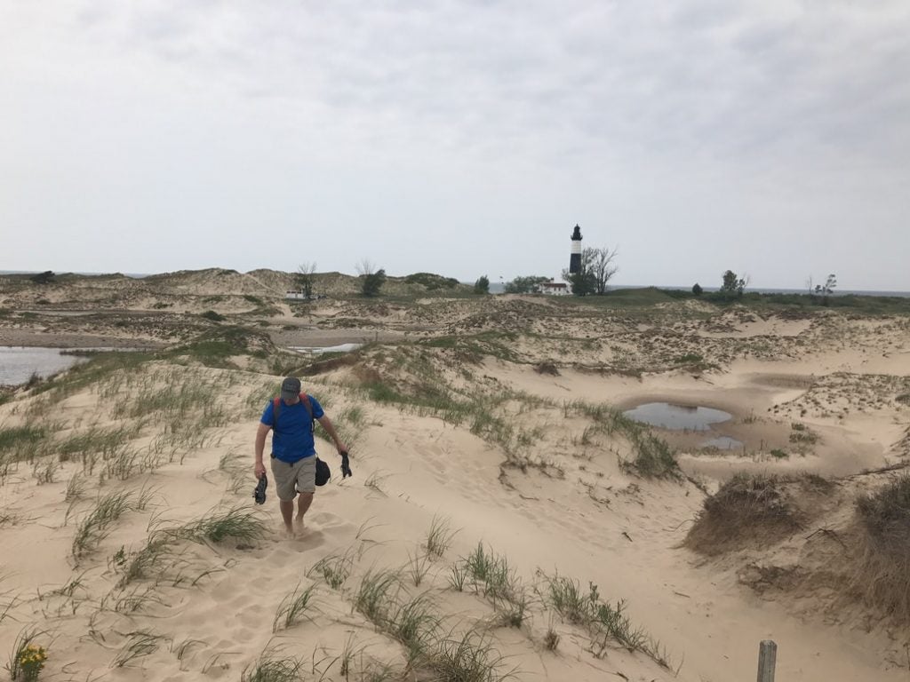 a man walks across sand dunes in front of a lighthouse near the lake