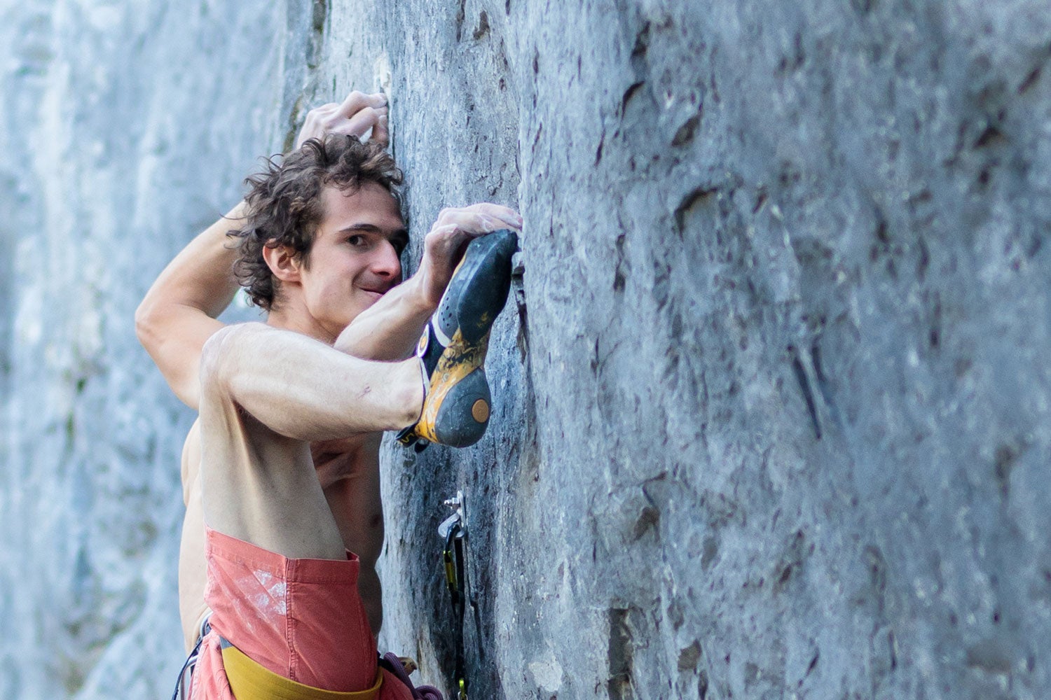 A climber clings to a vertical wall, grimacing as he matches he foot to his hand hold. 