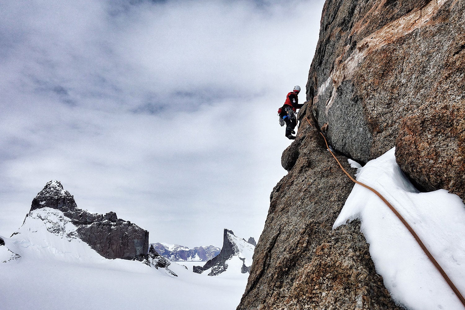 Rock climber perched on a rock face with snow in the background. 