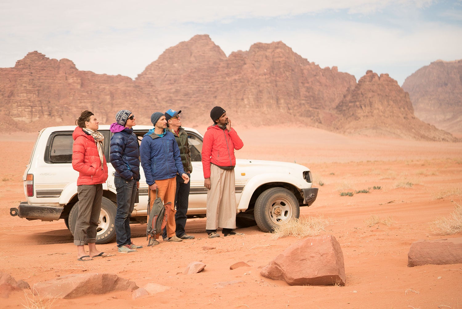 Five people standing in a red desert landscape next to a white SUV. 