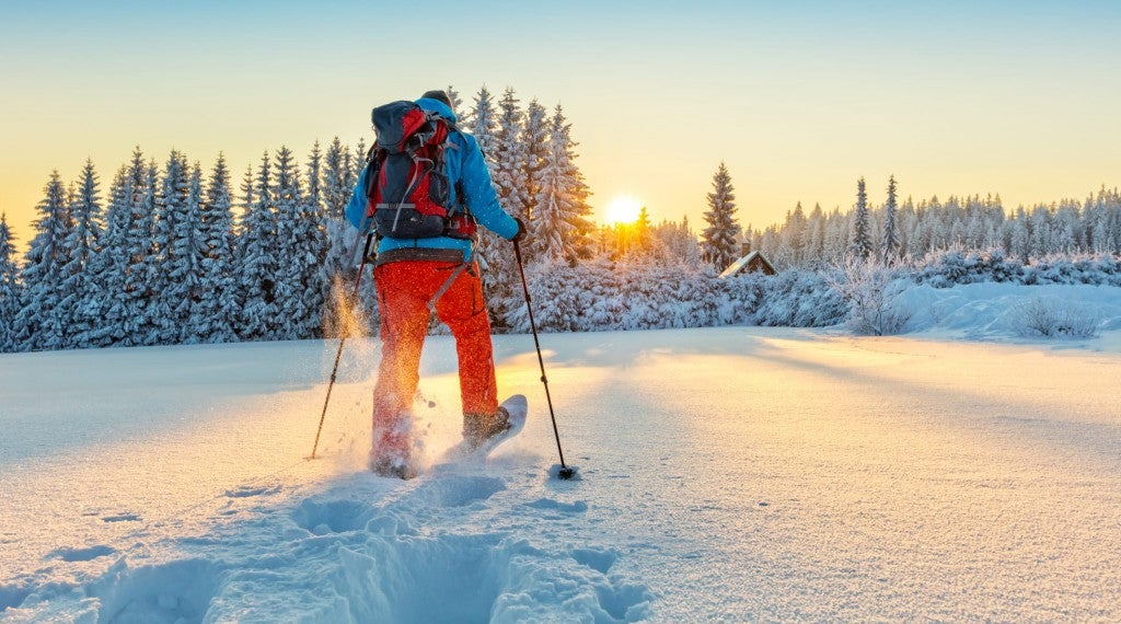 snowshoer in red pants and blue coat walking across snowfield towards sun and treeline