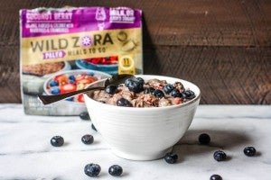 spoon with white bowl filled with oatmeal and blueberries with package of Wild Zora freeze dried meal in the background