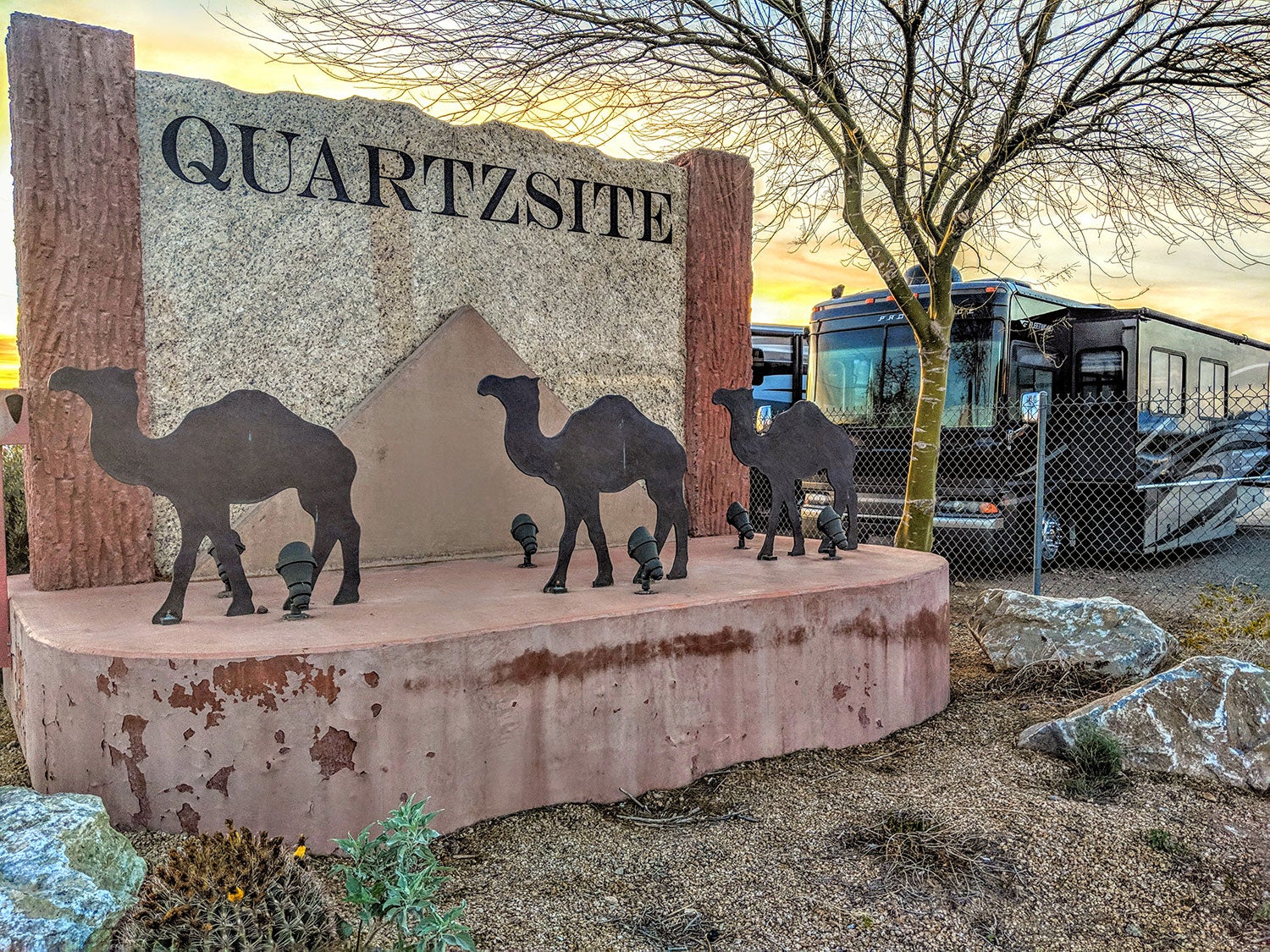 a sign made out of Quartzsite in arizona featuring three metal camels near a pyramid with the name of the town on it, next to a line of RV campers