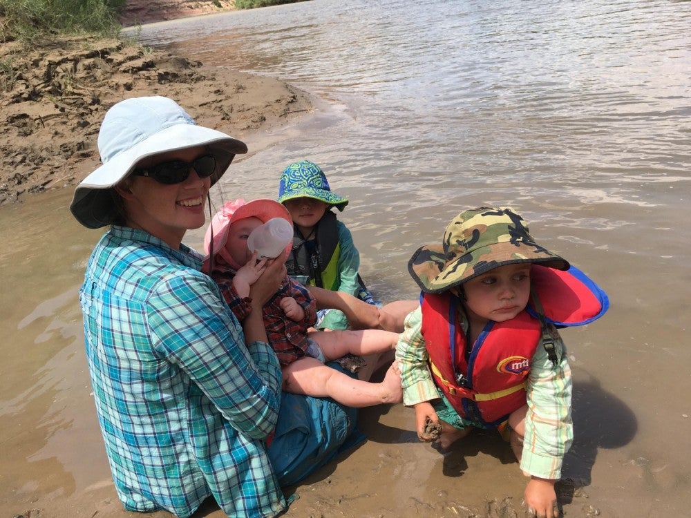 a mom bottle-feeding a baby near two toddlers sitting on a wet sandy beach