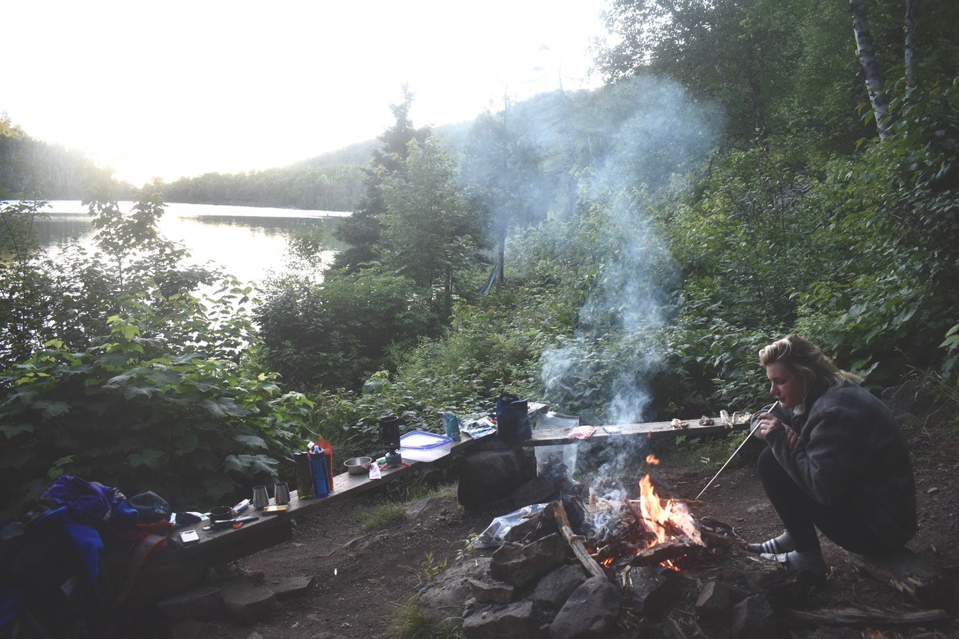 female camper pokes at campfire in the foreground with wooded lake in the background