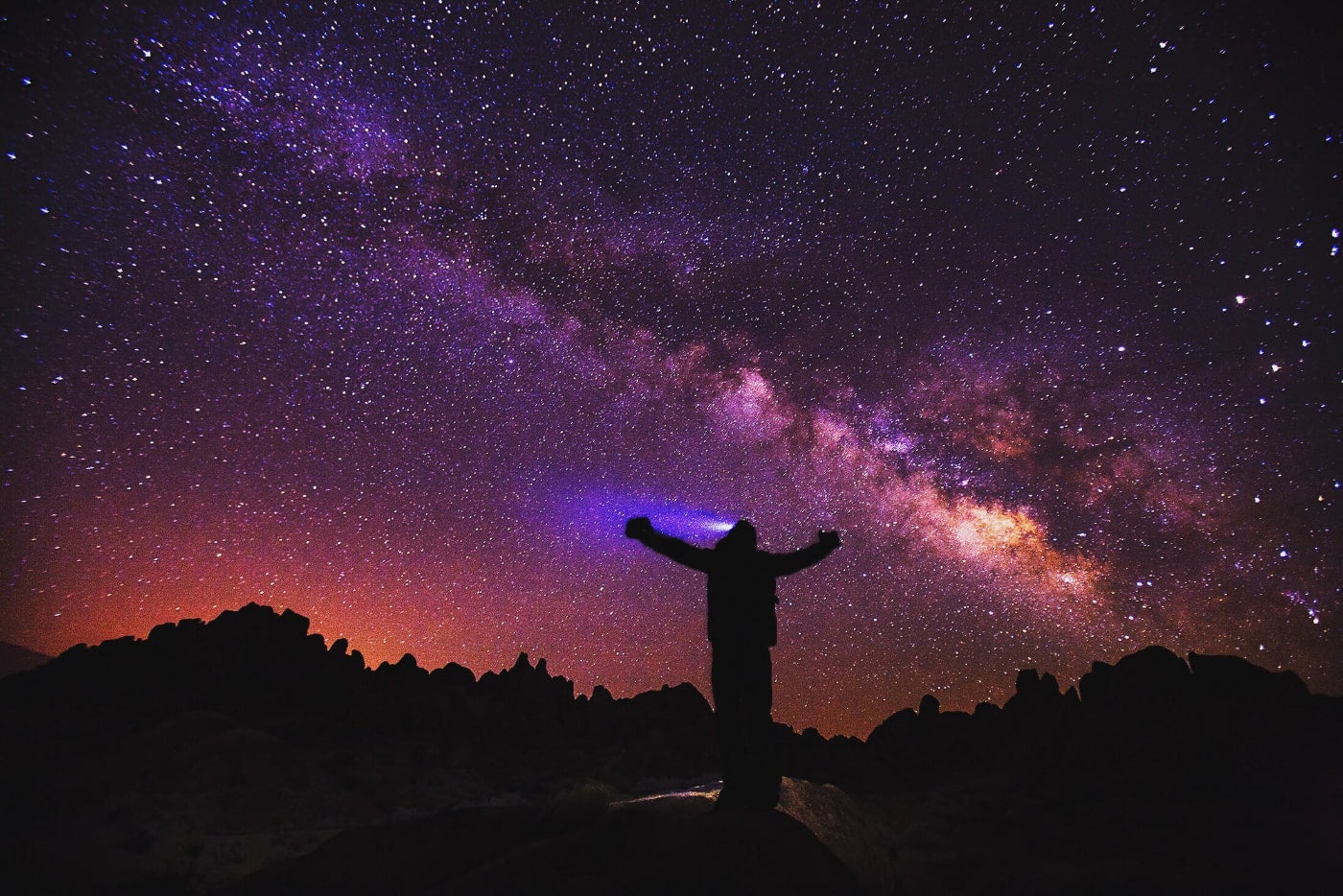 a silhouette of person with arms raised and a headlight beam shining on a silhouetted landscape of rocky hills and a purple night sky filled with stars.