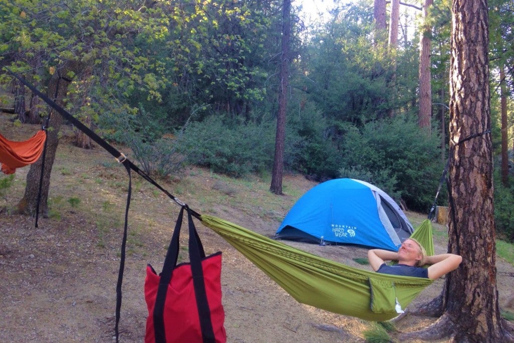 woman relaxing in green hammock at campsite with blue tent and tall pine trees in background