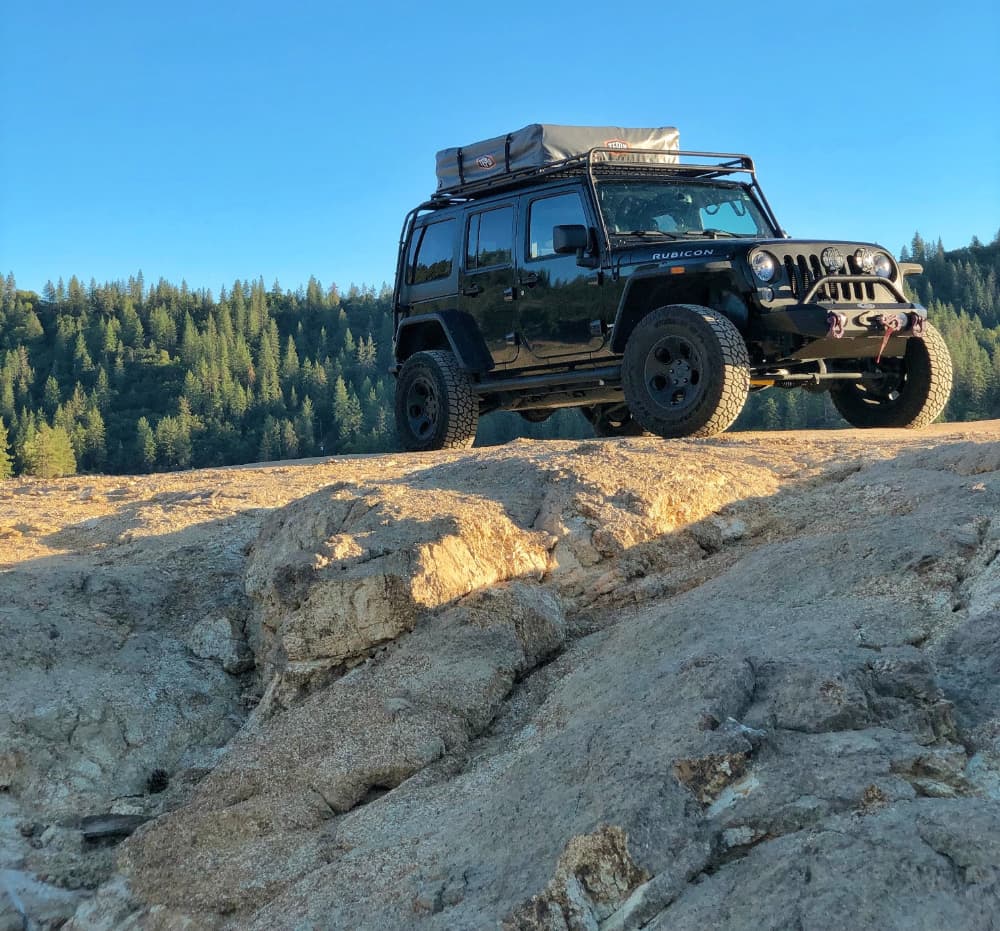 black jeep with rooftop tent parks on top of rocks with forest in background