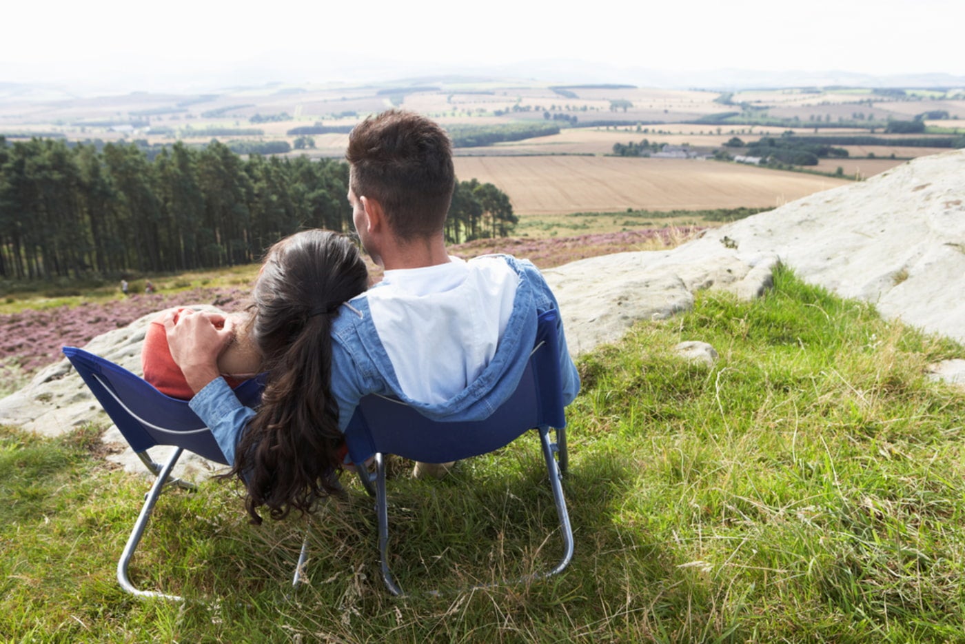 a couple sharing a blue loveseat camp chair, perched on a grassy hill and looking over an agricultural landscape.