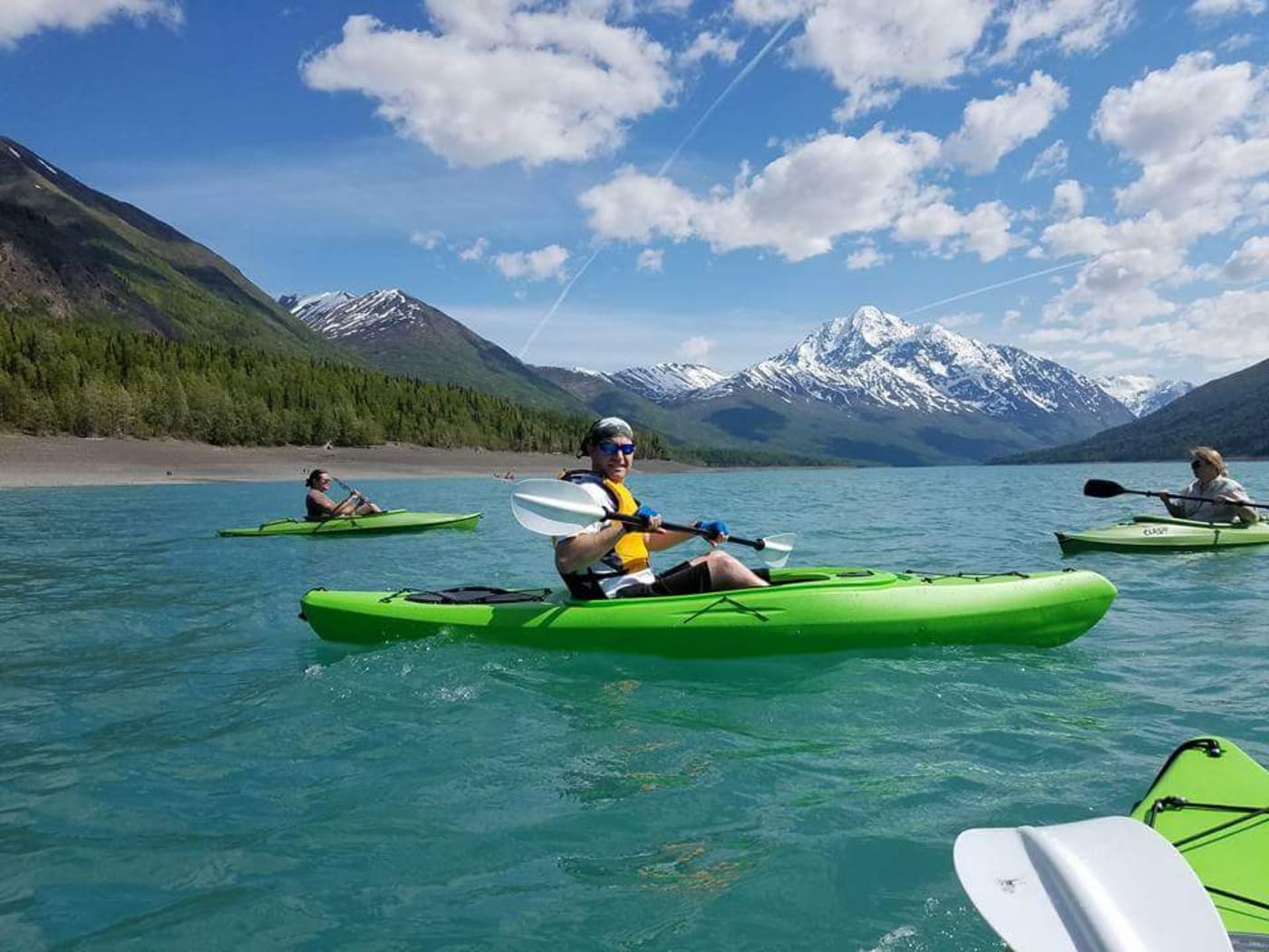 Kayakers on the glacial Eklutna Lake on a sunny day with snowcapped mountains in the distance.