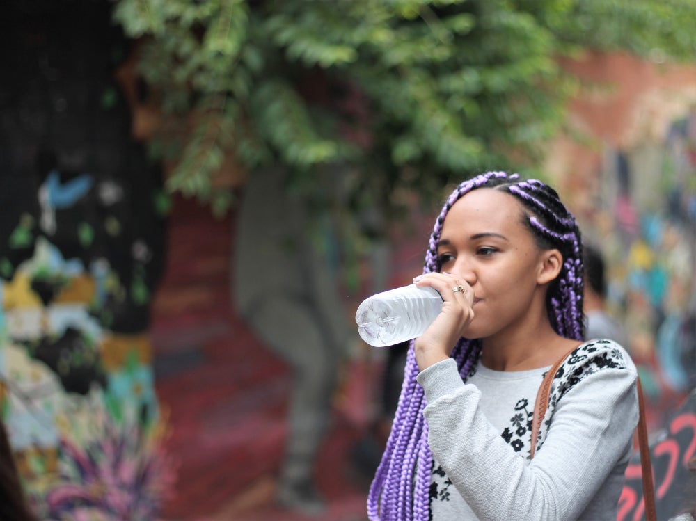 Woman wearing gray shirt and purple braids drinks water from water bottle, with trees and colorful walls in the background 