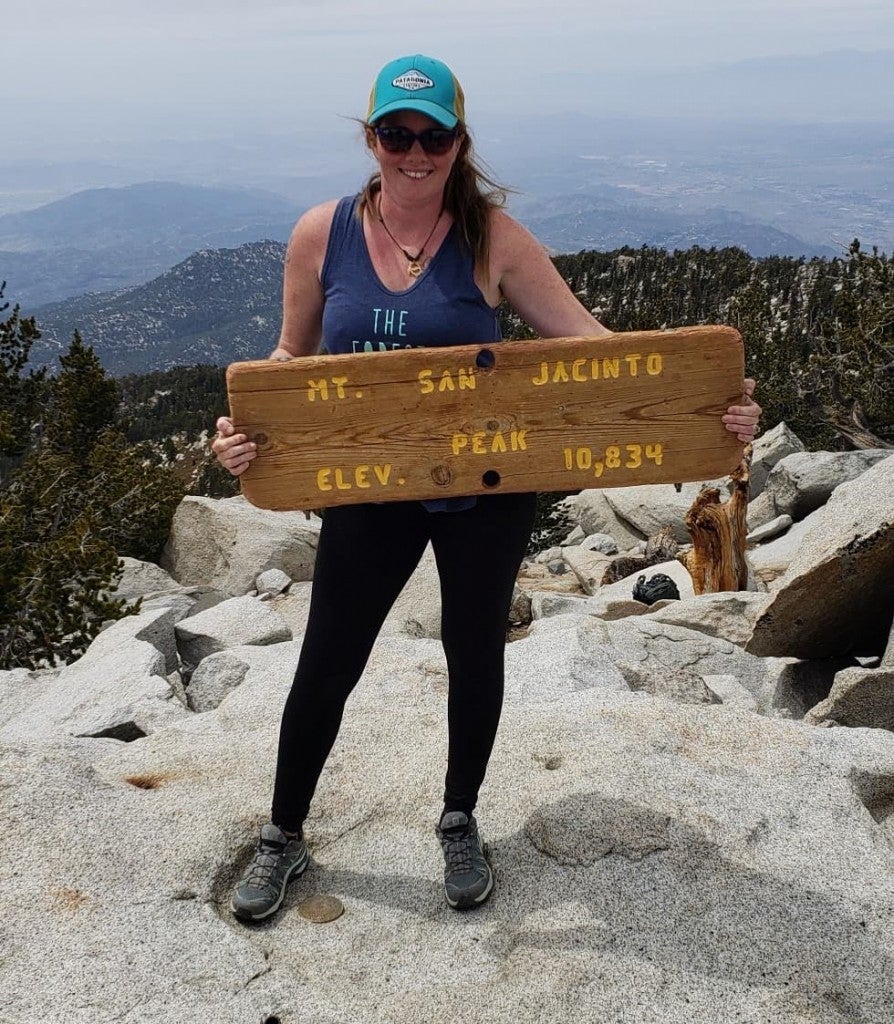 woman standing on top of mountain peak holding mt. san jacinto summit sign with smoky valley below