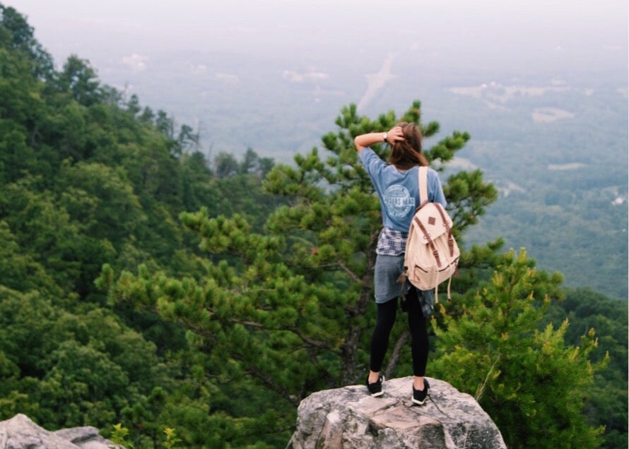 Woman wearing blue shirt and beige backpack overlooking a sweeping view of the forested valley surrounding Pilot Mountain State Park