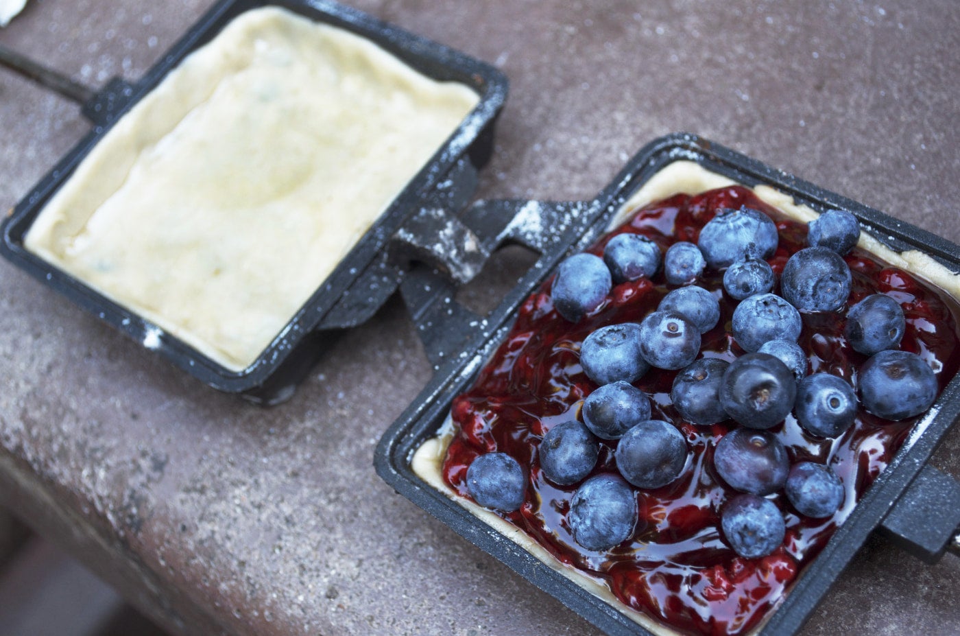 A cast iron double pie iron filled with dough and blueberry pie filling laid open prior to cooking.