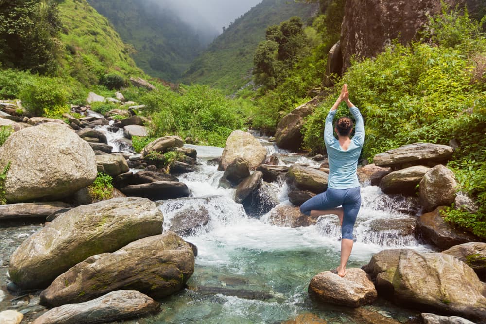 woman doing yoga pose standing on rock in mountain stream with misty mountains in the background