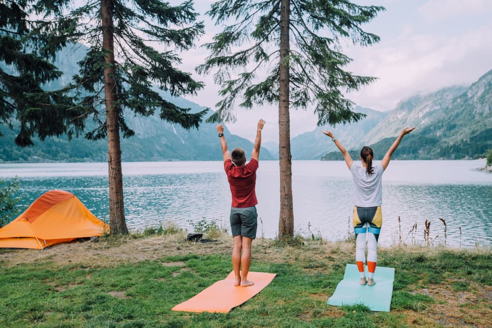 man and woman doing standing yoga pose next to orange tent overlooking mountain lake