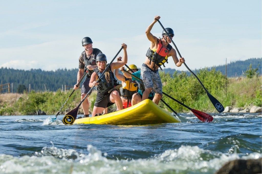 5 people on a large inflatable stand up paddle board racing down a river