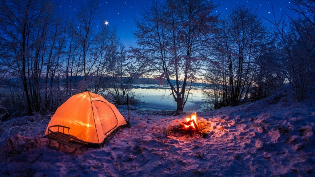 night shot of glowing orange tent and campfire in the snow on the edge of a partially frozen lake