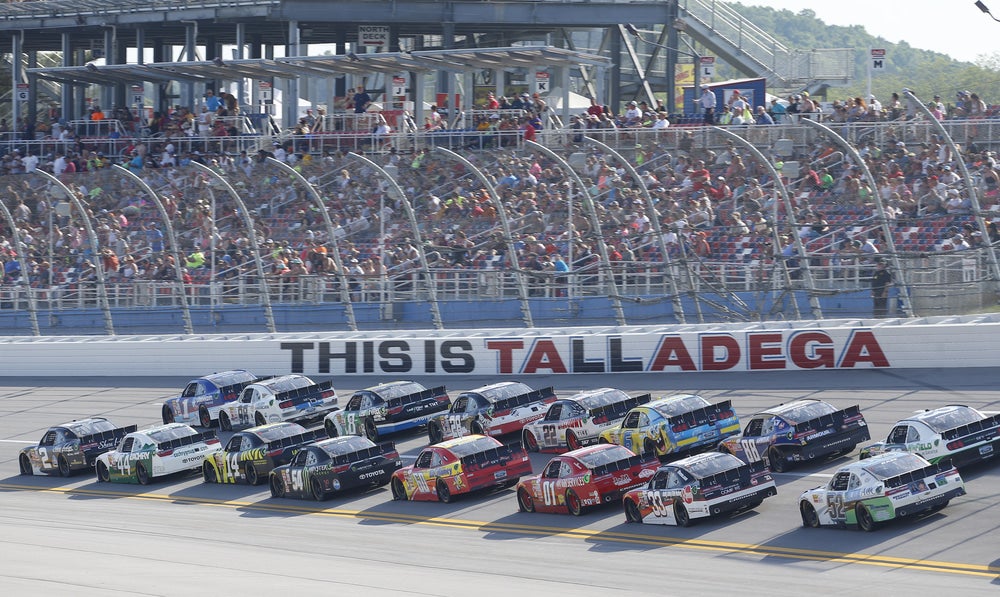 Multiple cars close together racing on NASCAR home track in Alabama. Sign on track says "This is Talladega"