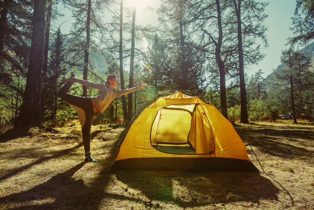 woman doing standing yoga pose next to yellow tent in the forest