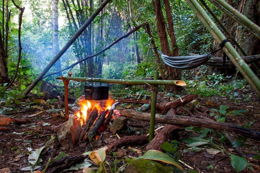 a hammock hangs in the jungle beside a lean-to cooking campfire