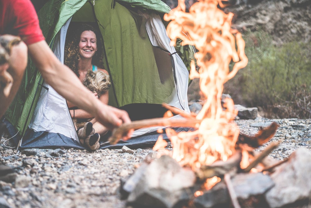 A woman sitting in a tent holding a dog near a person lighting a fire