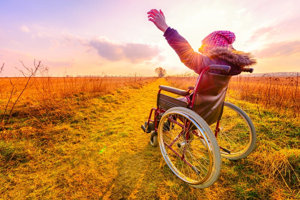 Woman with pink hat in a wheelchair holding up her arms in a celebratory fashion at sunset