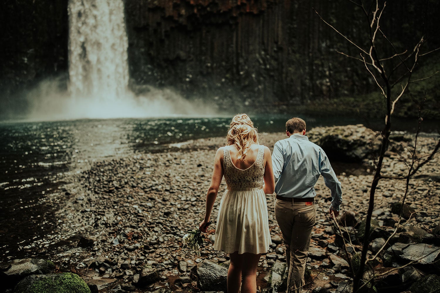 a man and a woman walking toward a rocky island in a lake at the end of a tall waterfall