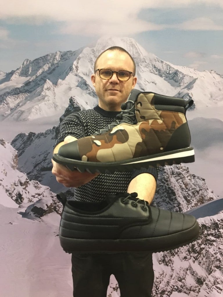 a man holds two puffy slippers from holden outerwear in front of a mountain background