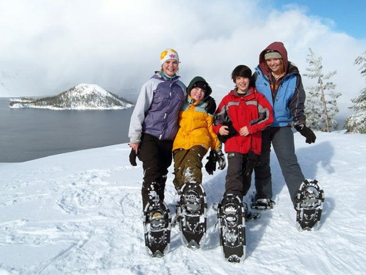 Two women and two boys wearing snowshoes pose together in front of crater lake in the winter.