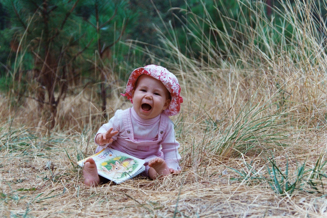 baby girl with pink hat and overalls sitting in tall grass and laughing