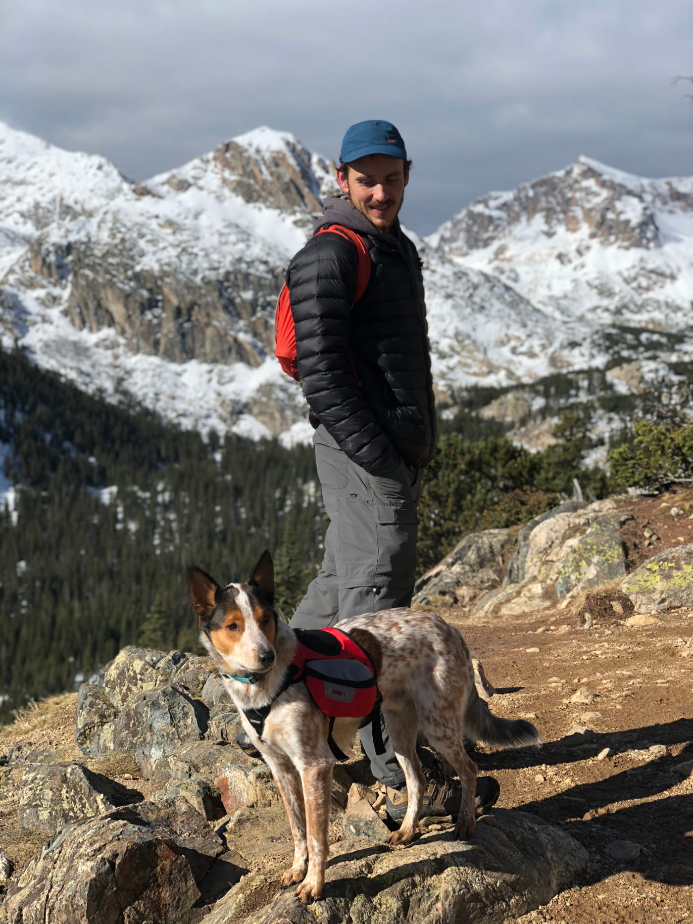 a man and a dog stand on the ridge of a mountain hiking trail in front of a snowy mountain range