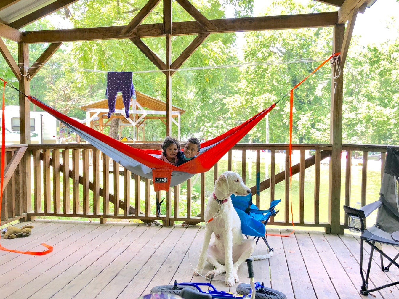 two young children smiling from red hammock hung on deck as white dog sits in the foreground