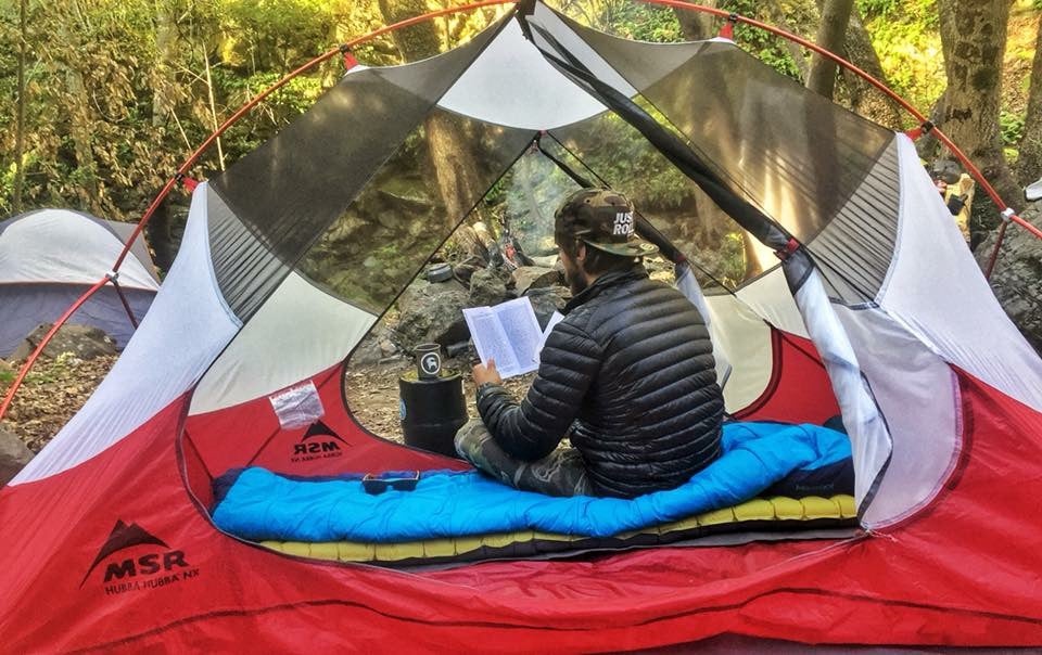 Guy with a brown hat facing away from the camera sitting in a tent reading with a black puffer jacket on a blue sleeping bag on a green sleeping m,at in a red and white MSR tent.