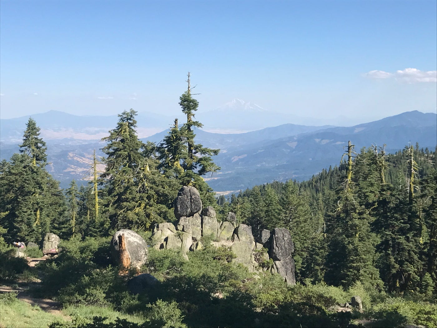 A stack of rocks on a hill covered in evergreens, in the distance is faint outline of Mount Shasta in southern oregon