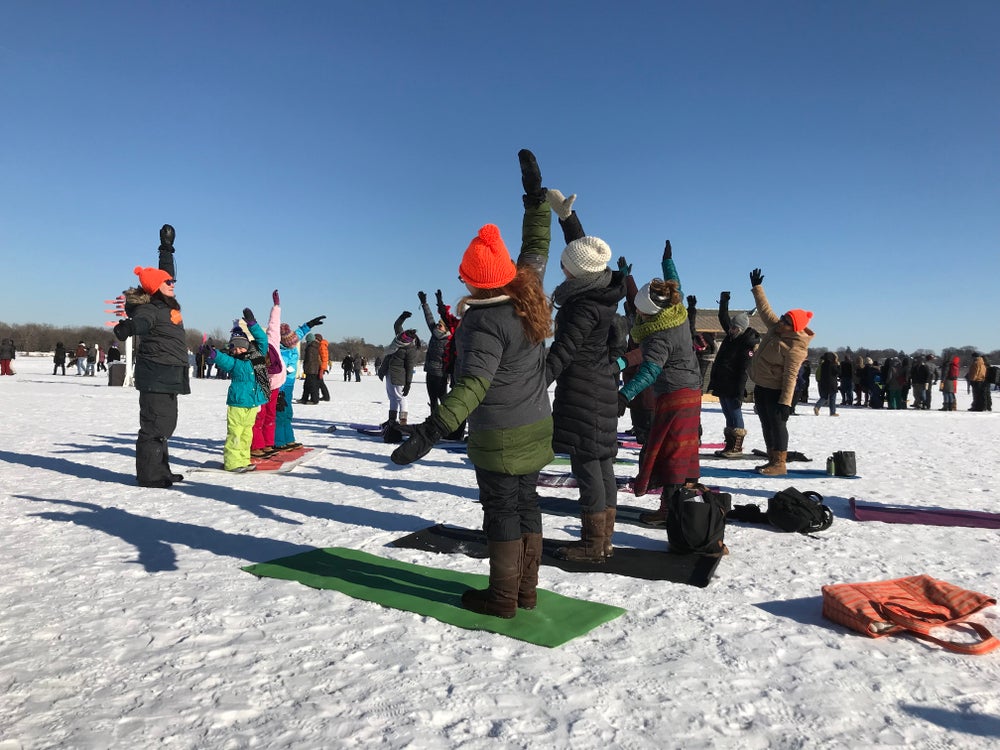 people in winter apparel stretch their right arms over their heads as they participate in outdoor snow yoga