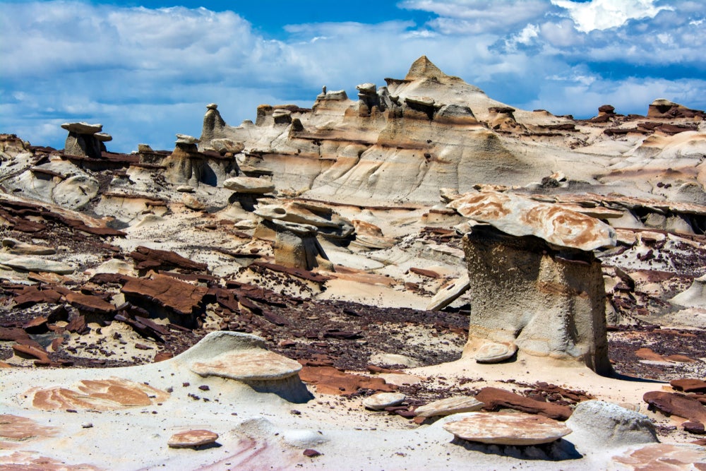 White and dark brown straited rock formations in the Bisti Badlands against a cloudy bright blue sky.