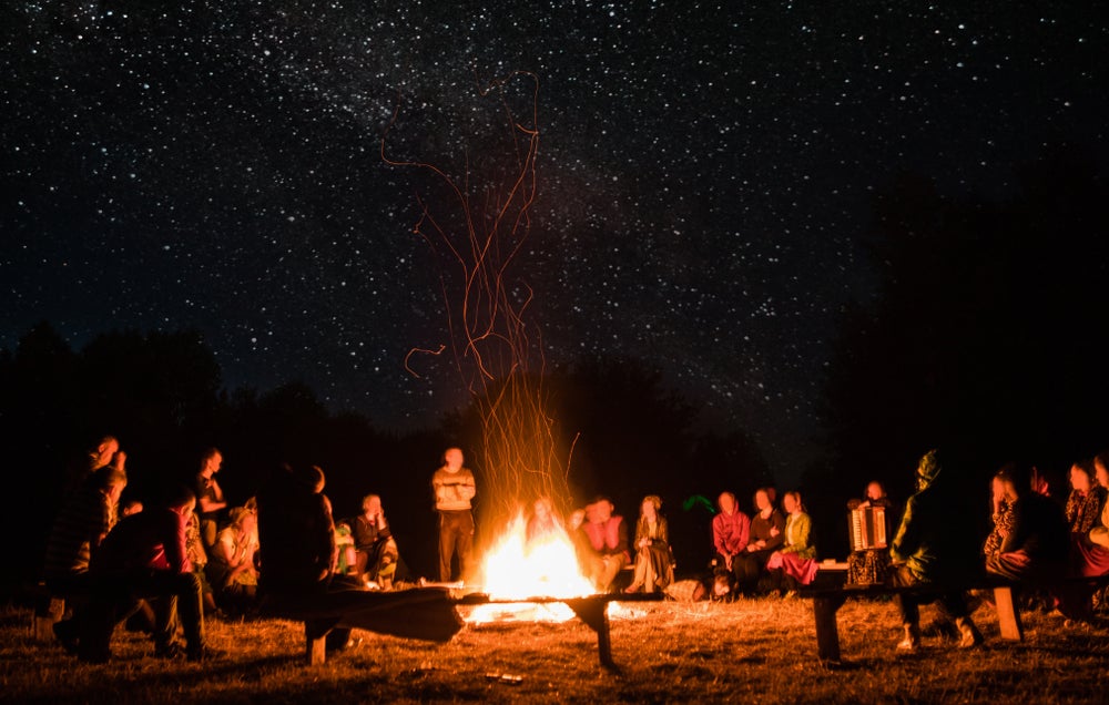 Large group sits on wooden benches around a campfire at night.
