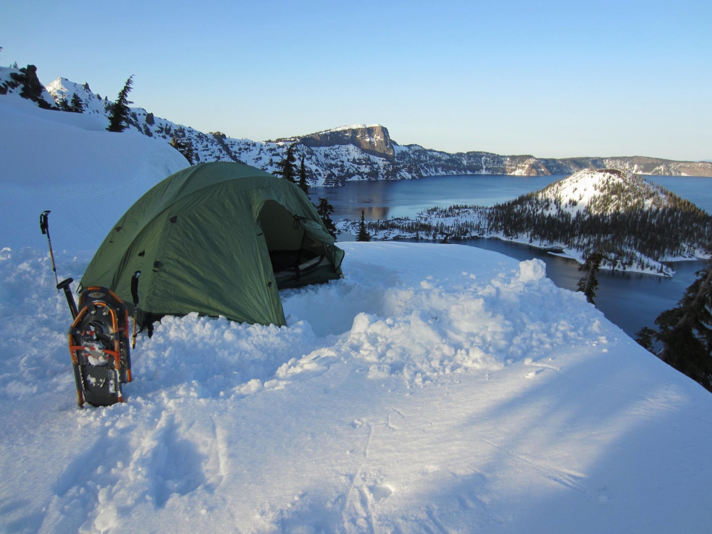 Orange snowshoes and a green tent sit in the deep snow beside crater lake in winter.