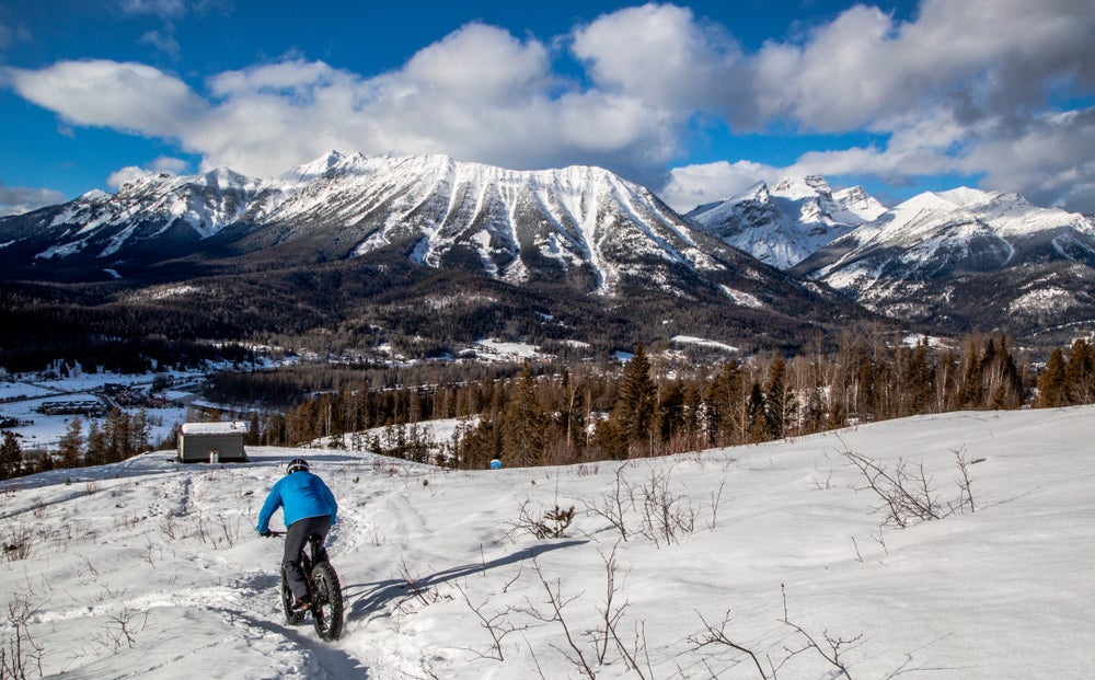 Person in winter gear riding a fat bike through a snow field towards a mountain range