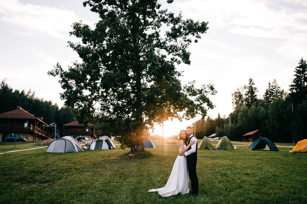 Couple stands together in formal wedding attire, in front of large trees, a lodge building, and a cluster of dispersed tents.