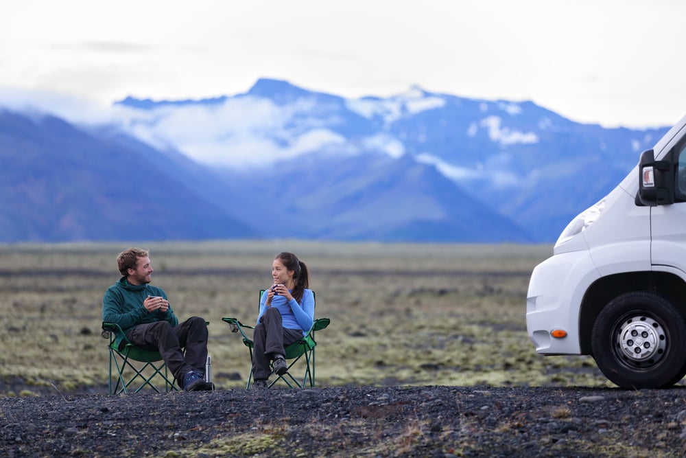 Couple sits in cozy sweaters in camp chairs in a field beside their RV drinking coffee.